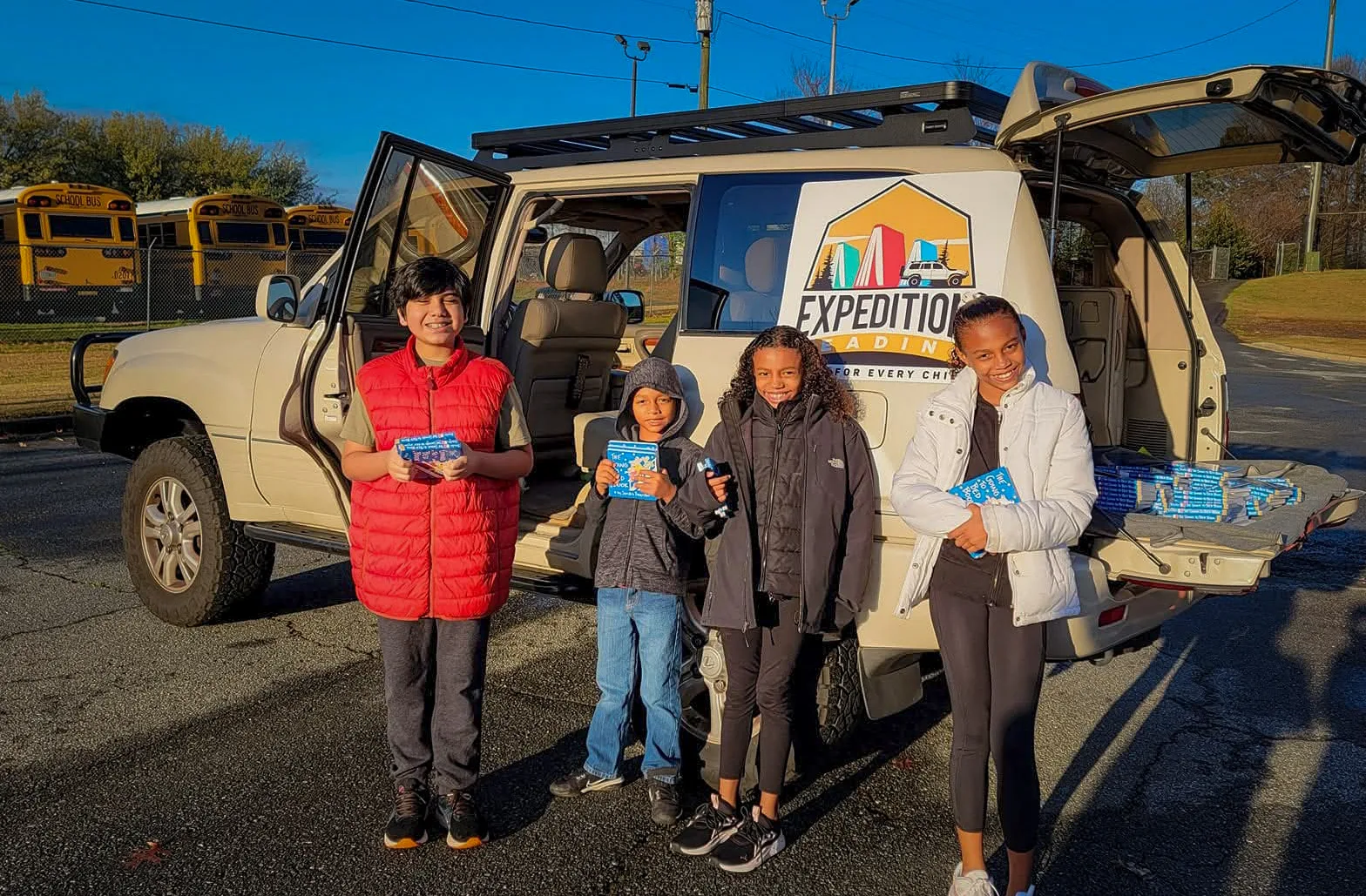 Four children stand beside the Expedition Reading delivery vehicle on a sunny afternoon, each holding a book they just received